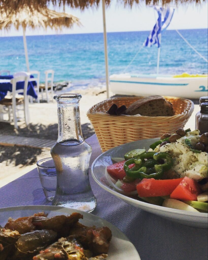 a plate of dolmadakia and a plate of Cretan salad on a table with a basket of fresh bread, Cretan raki tsikoudia and a boat in the background representing Cretan hospitality in Lasithi, Crete.