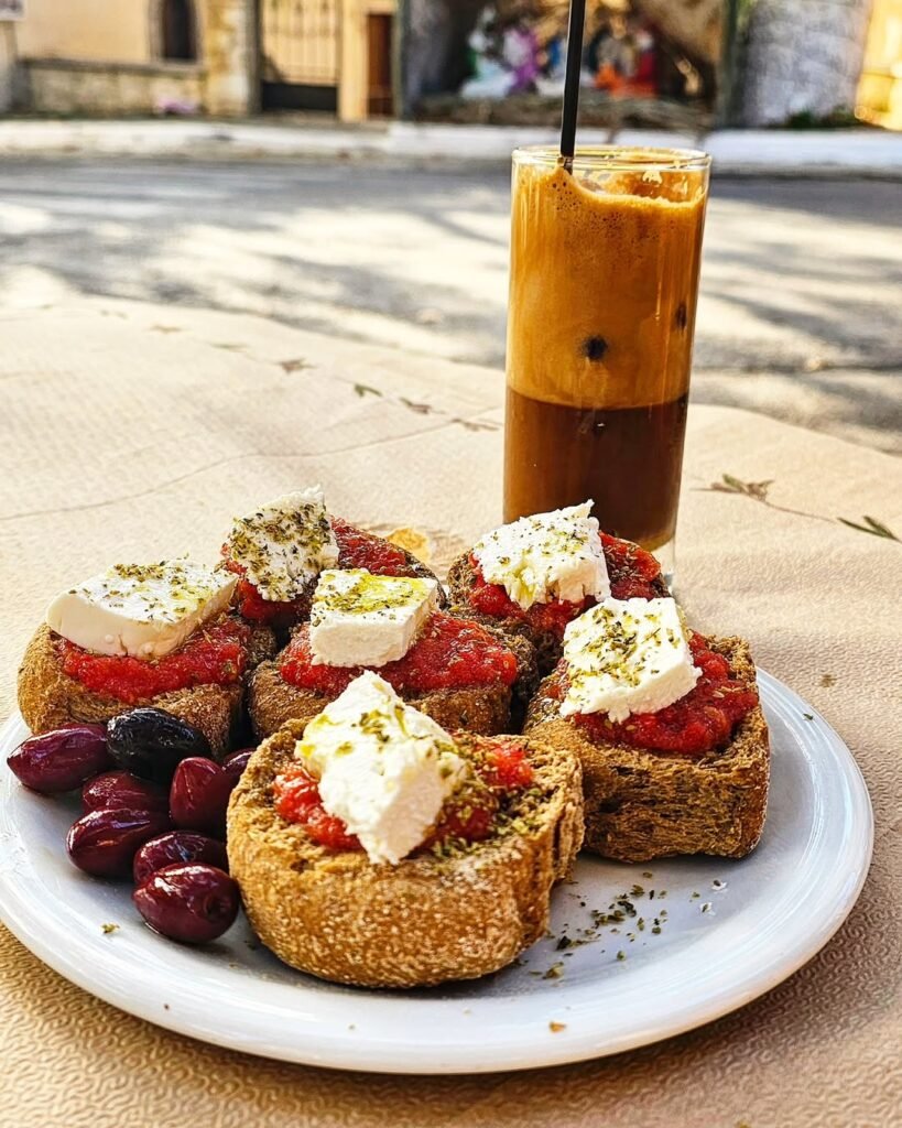 A classic taste of Crete—dakos topped with fresh tomato, creamy mizithra cheese, olive oil, and oregano, enjoyed with a frappe in the beautiful atmosphere at To Kafeneio Michael in Armenoi, Chania. 