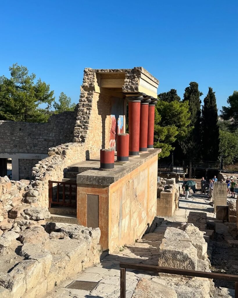 Visitors explore the famous North Entrance of Knossos Palace with its distinctive red columns and partially restored frescoes, where over one million tourists experienced Minoan history in 2024 before the upcoming ticket price increase to €20 in April 2025.