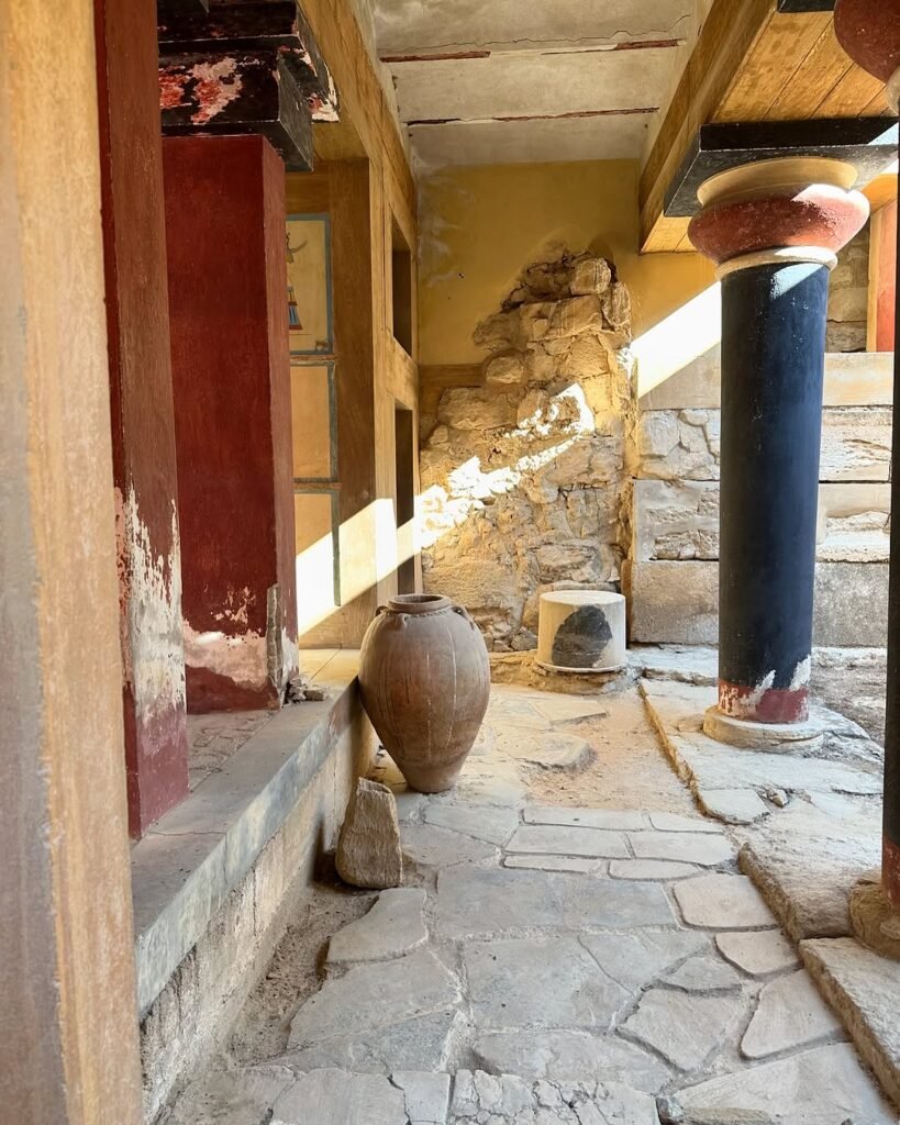 Interior passageway at Knossos Palace featuring colorful columns, ancient stonework, and storage vessels that will benefit from the €3.5 million modernization plan aimed at preserving these 4,000-year-old architectural elements while improving visitor access.