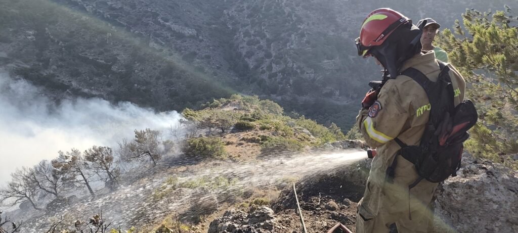 A firefighter continues monitoring operations in southeastern Crete on July 4th, 2025, as smoke still rises from smoldering areas. The main fire has been extinguished, but crews remain vigilant for potential flare-ups. 
