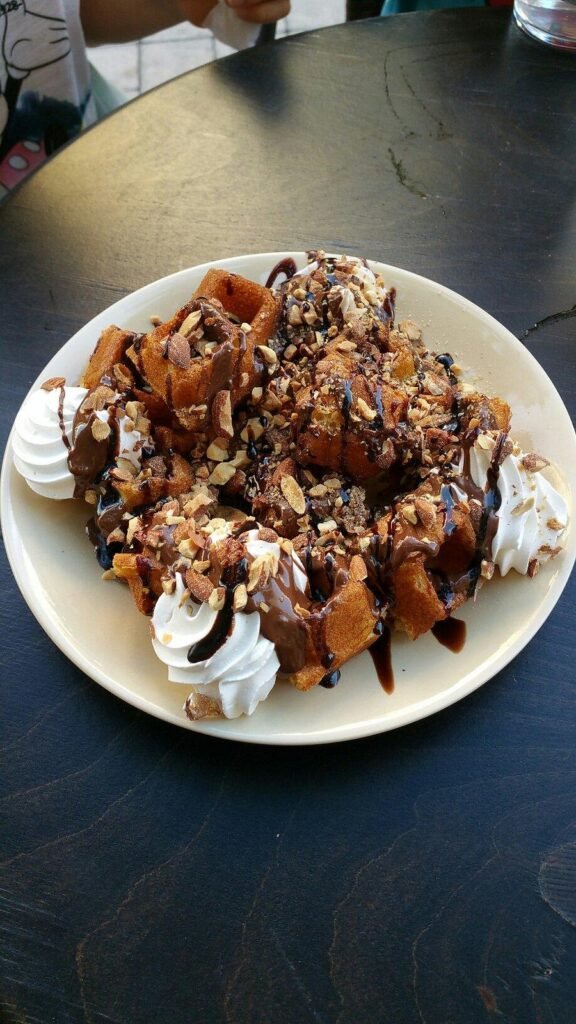 Decadent waffles at Karakatsanis in Paleochora piled high with chocolate sauce, crushed nuts, whipped cream, and chocolate drizzle, showcasing the generous portions that have made this dessert institution a beloved destination in southwest Crete. Photo Credit: Melania T.