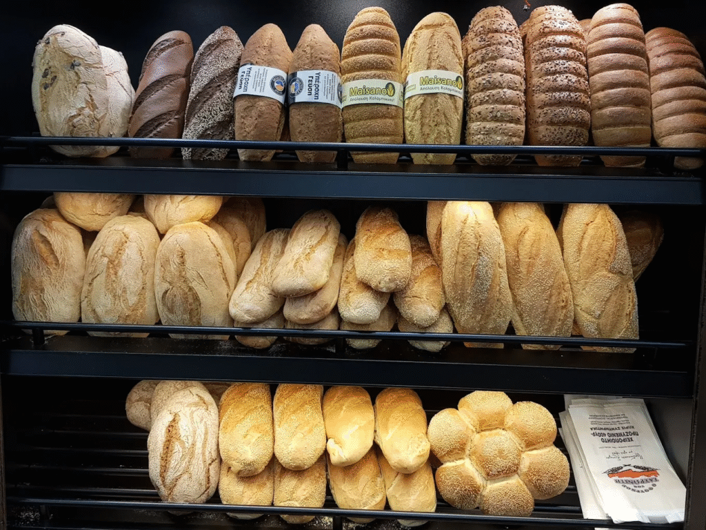 Freshly baked artisan breads displayed at Papanikolakis Bakery in Chania, featuring rustic sourdough loaves, sesame-topped braided breads, and traditional baguettes. Photo Credit: Papanikolakis Bakery